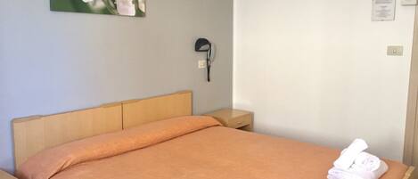 A neatly made bed with an orange bedspread and two folded white towels. A small bedside table is visible to the left of the bed.