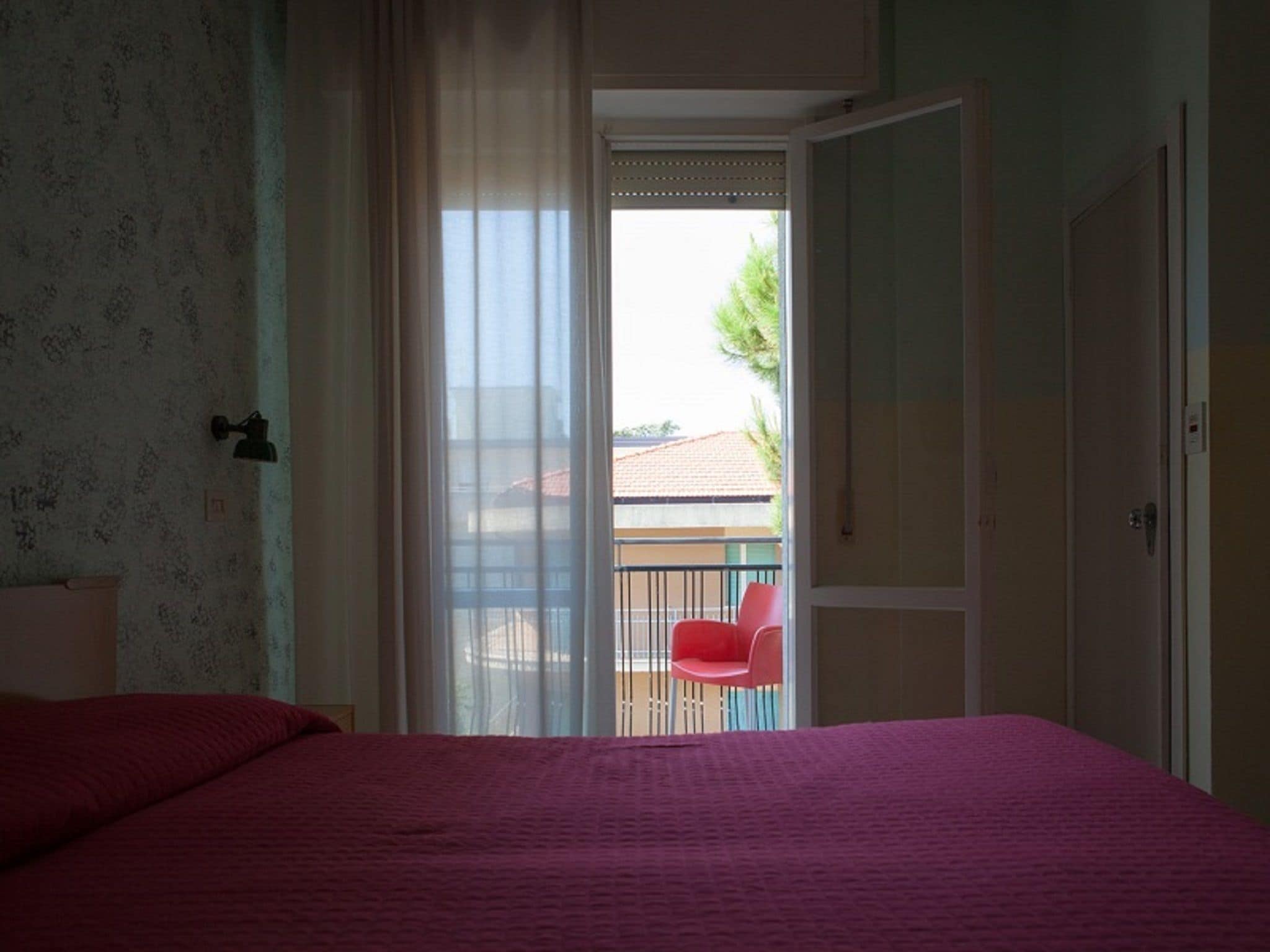 A bedroom with a bed, a window with curtains, a balcony with a chair, and a door.