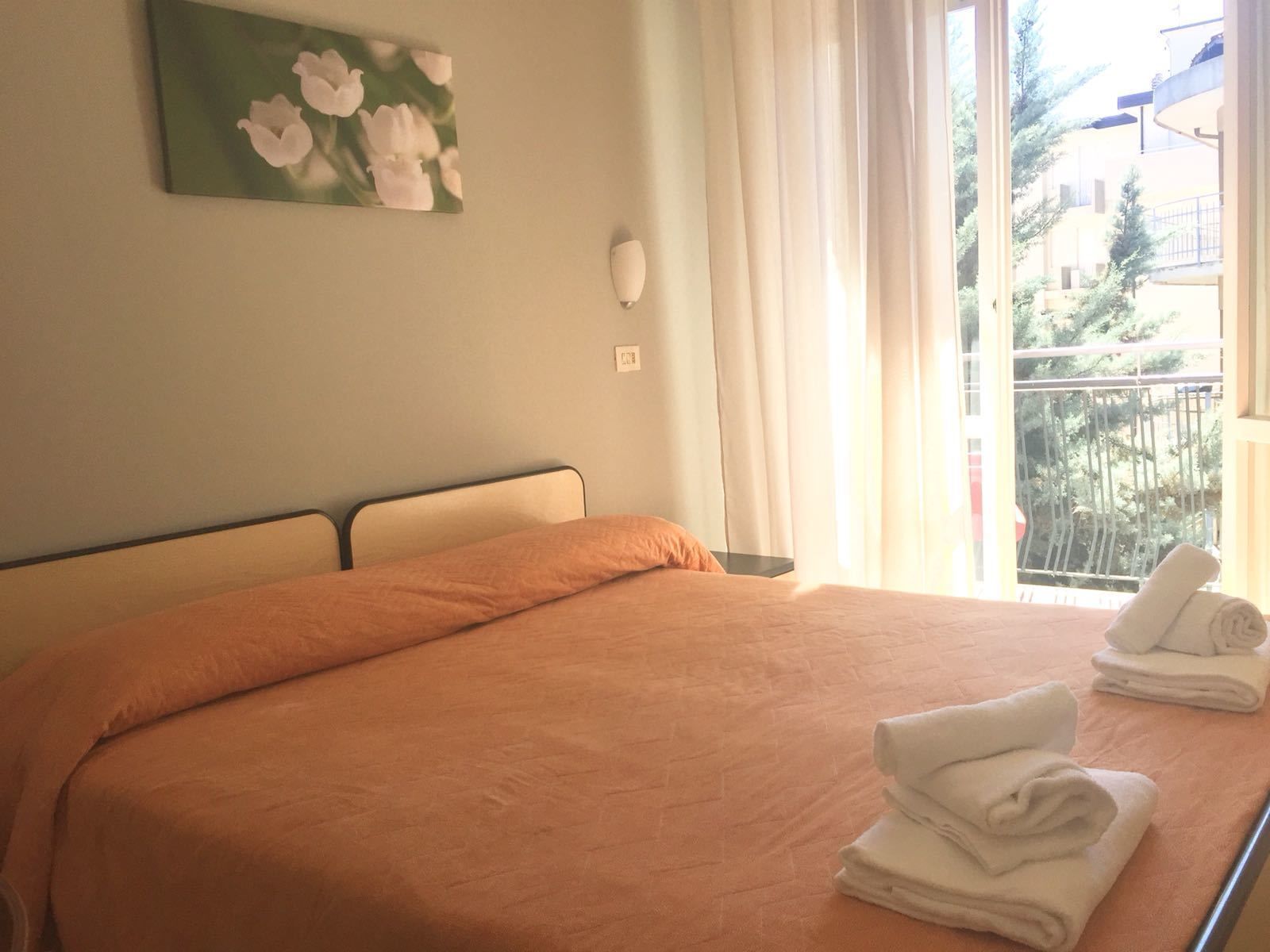 A hotel room with a bed, a window with curtains, a painting on the wall, and a stack of towels on the bed.