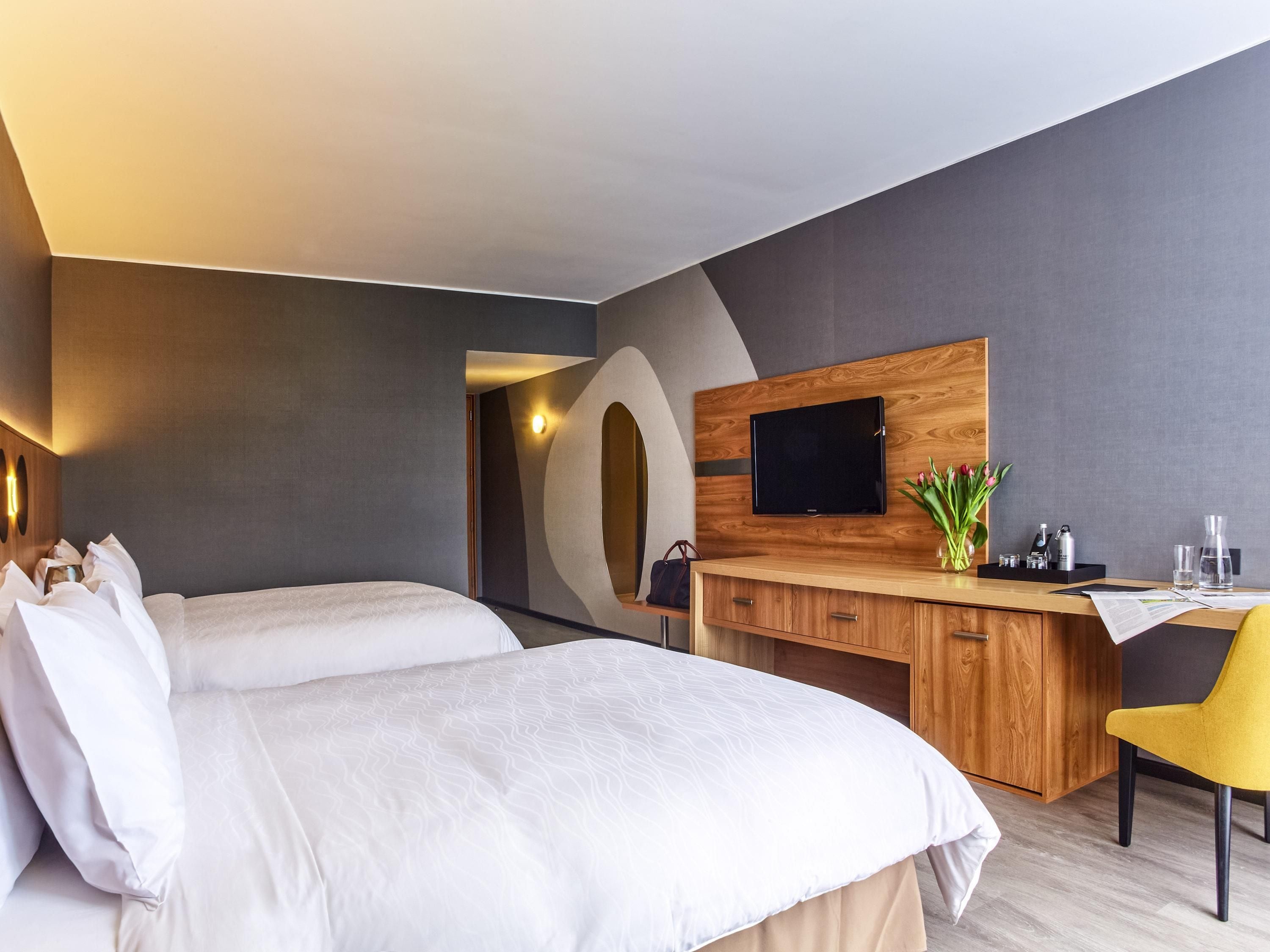 A modern hotel room with two beds, a wooden desk, a flat-screen TV, and a small desk lamp.