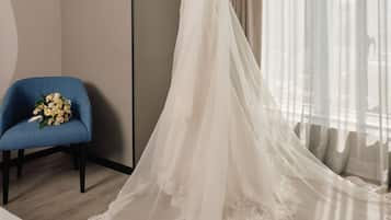 Um vestido de noiva em um manequim em um quarto com uma cadeira azul, um buquê e uma grande janela com cortinas leves.