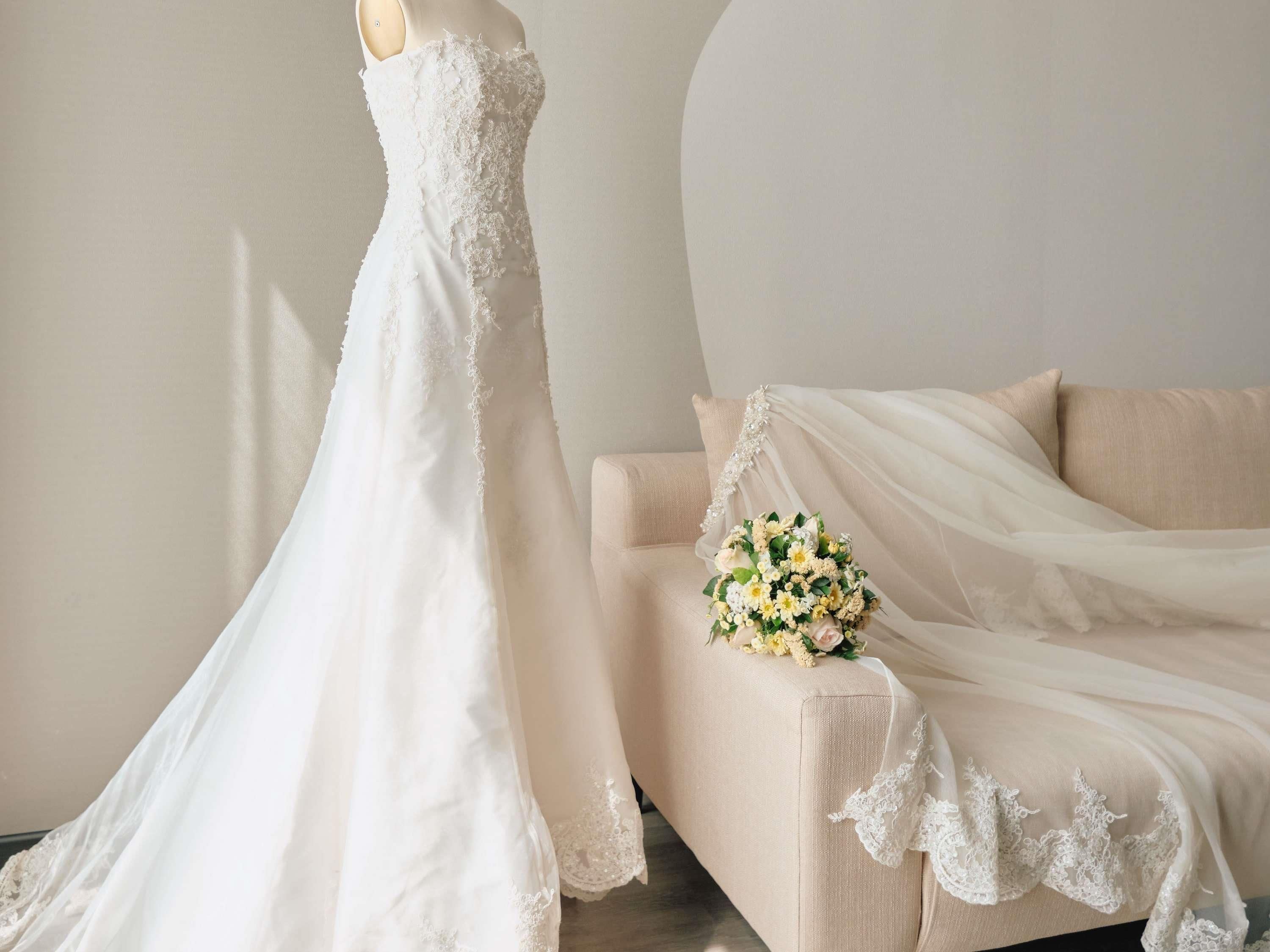 Un maniquí con un vestido de novia blanco con detalles de encaje, un sofá beige con un ramo de flores y cortinas traslúcidas.