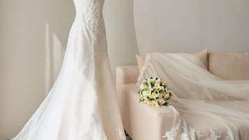 Un maniquí con un vestido de novia blanco con detalles de encaje, un sofá beige con un ramo de flores y cortinas traslúcidas.