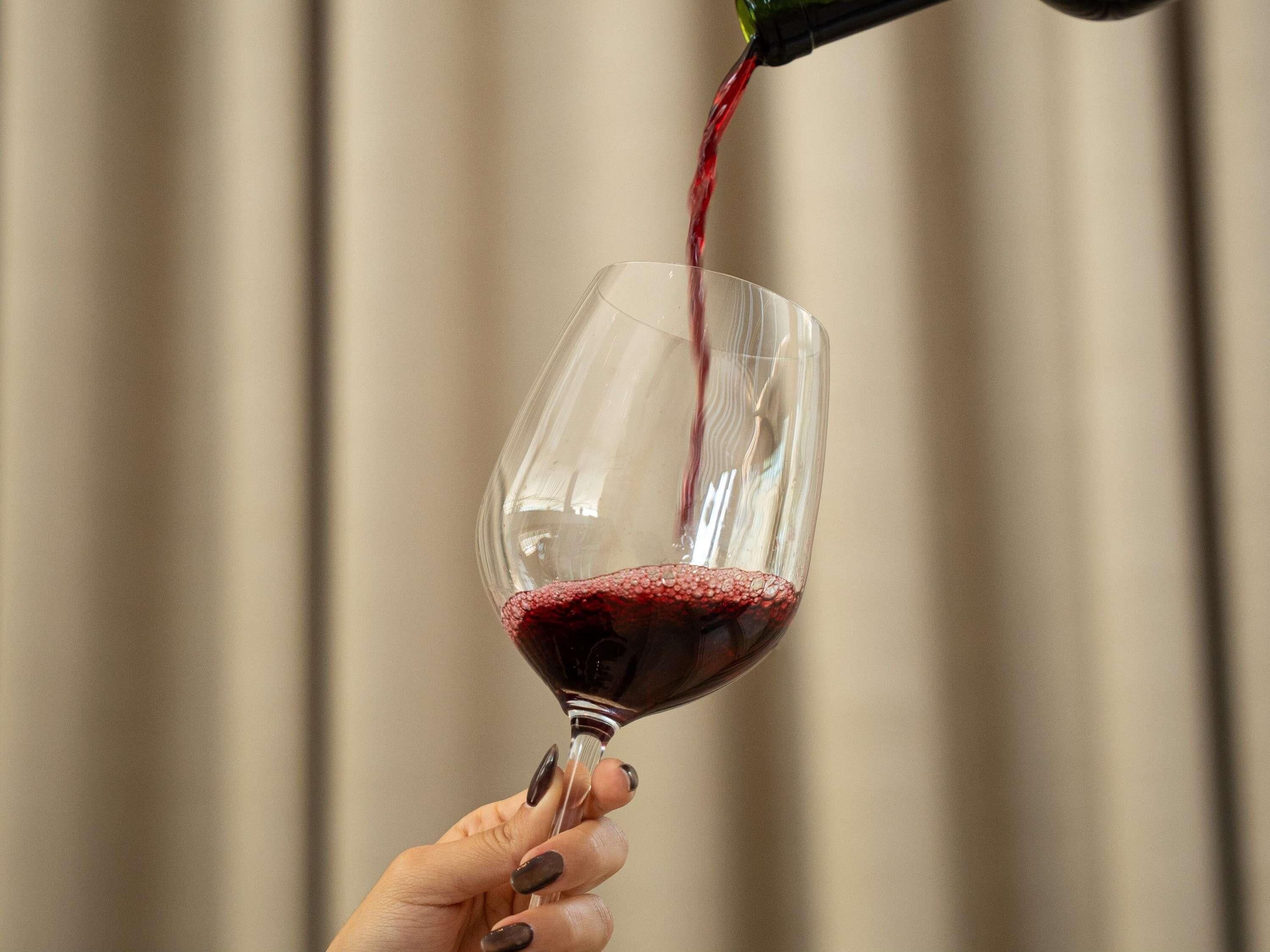 A hand holding a wine glass with red wine being poured into it.
