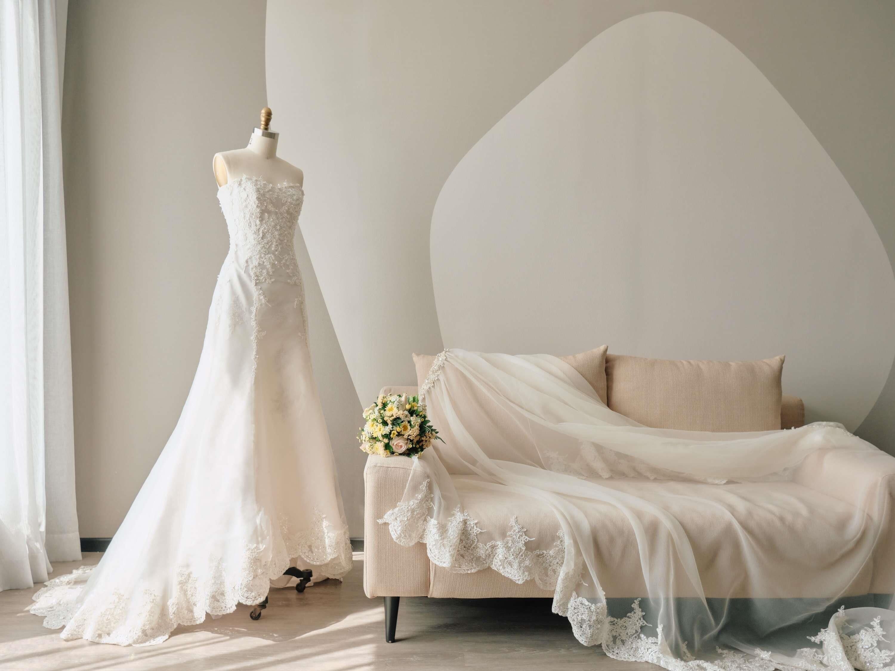 Une pièce où se trouve un mannequin portant une robe de mariée, un lit recouvert d’un voile transparent et un bouquet de fleurs posé sur le lit.