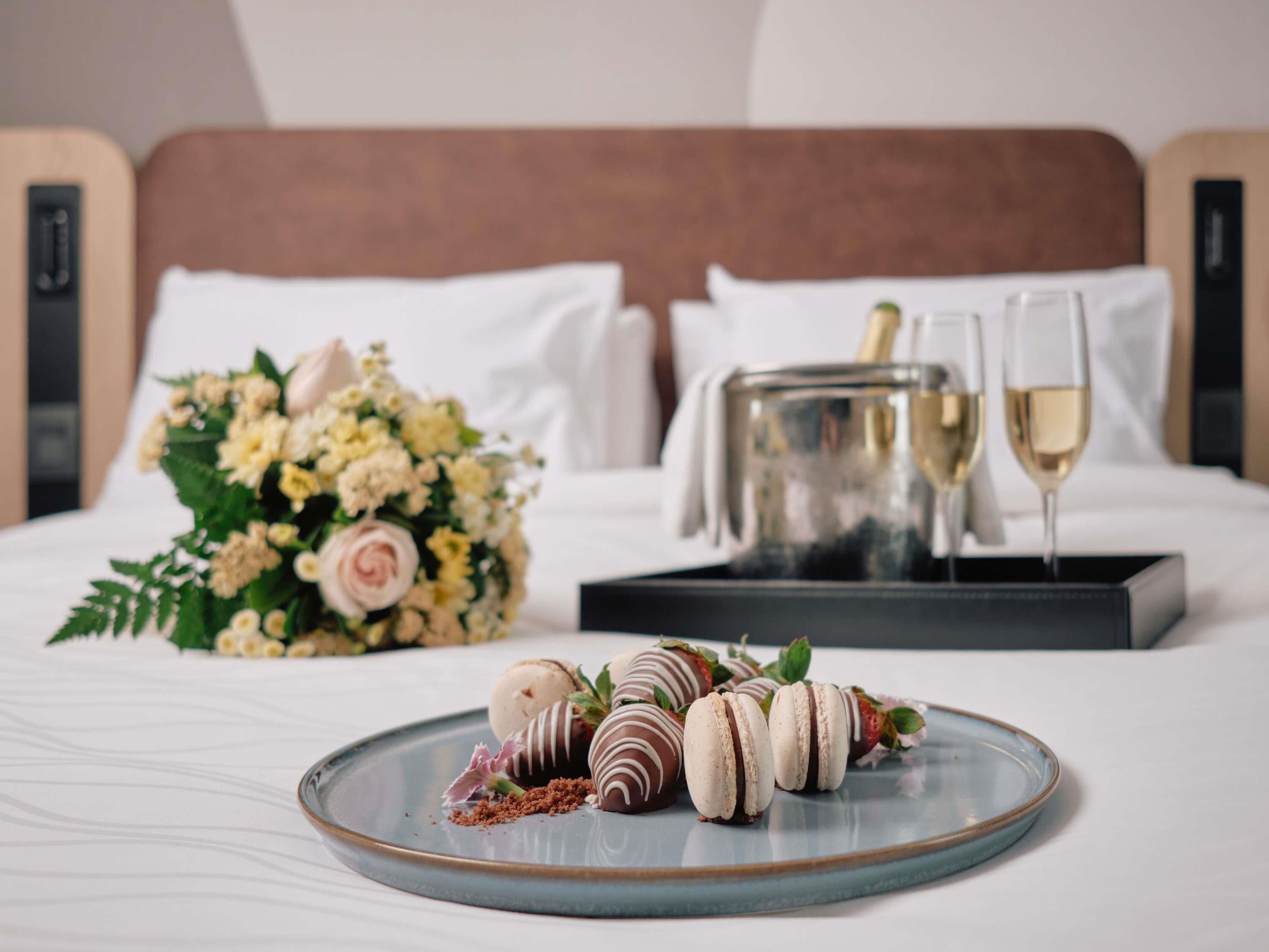 A bed with a bouquet of flowers, a tray with two glasses of wine, and chocolate-covered strawberries and macarons on a plate.