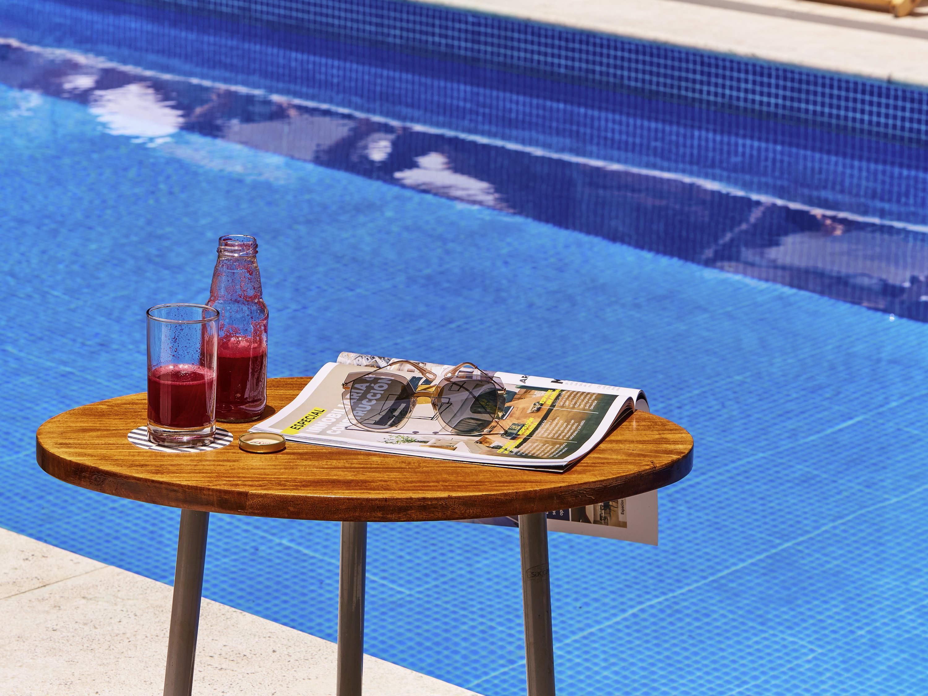 Ett träbord med en flaska, ett glas och ett par solglasögon, placerat på en tidning bredvid en pool.