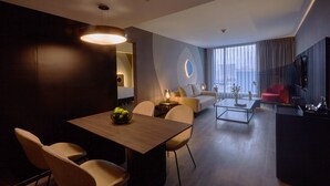 A modern hotel room with a dining area, a living room with a sofa and armchairs, a flat-screen TV, and a large window with curtains. - Pullman Lima San Isidro (Lima)