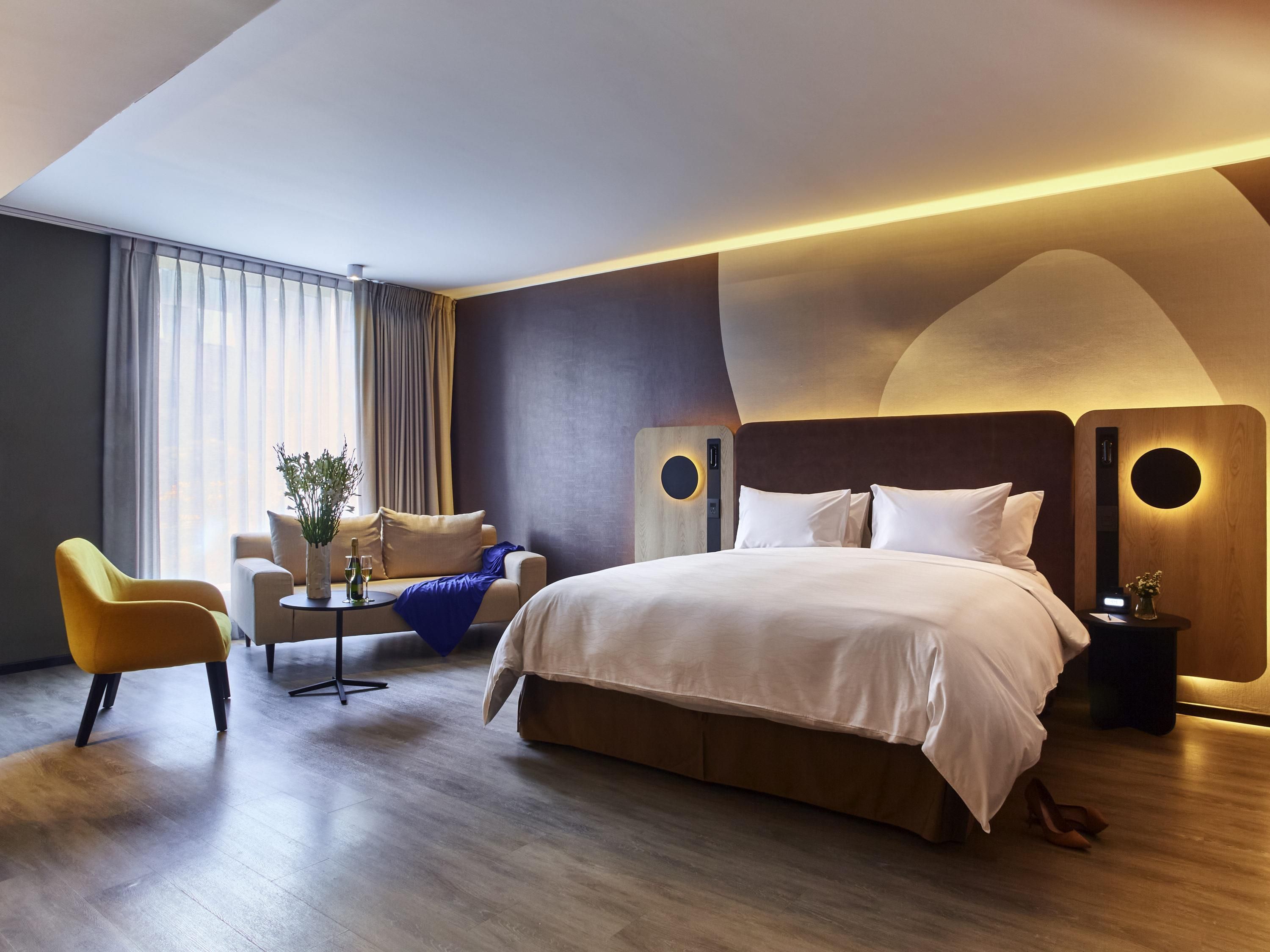 A modern hotel room with a large bed, a sofa, a small table with a vase and a bottle, and a view of the outdoors through the window.