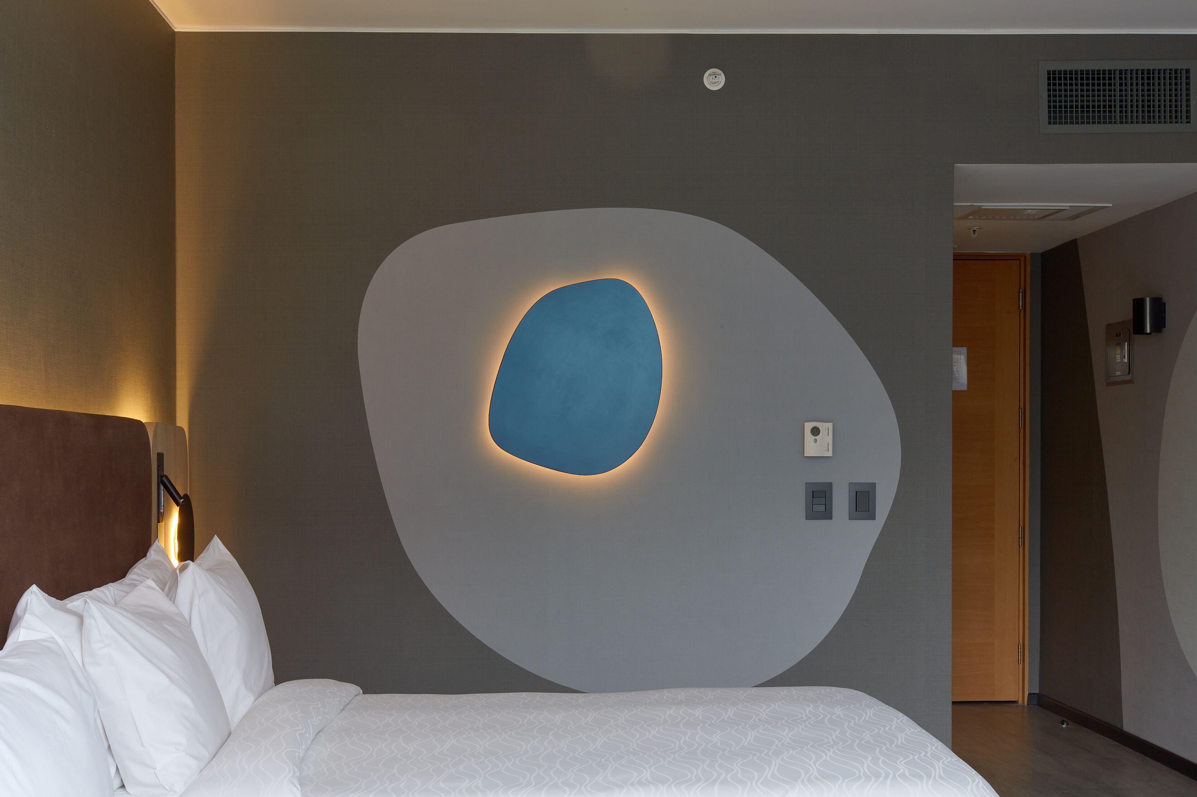A modern hotel room with a large bed, a wall-mounted light fixture, and a door leading to another room.