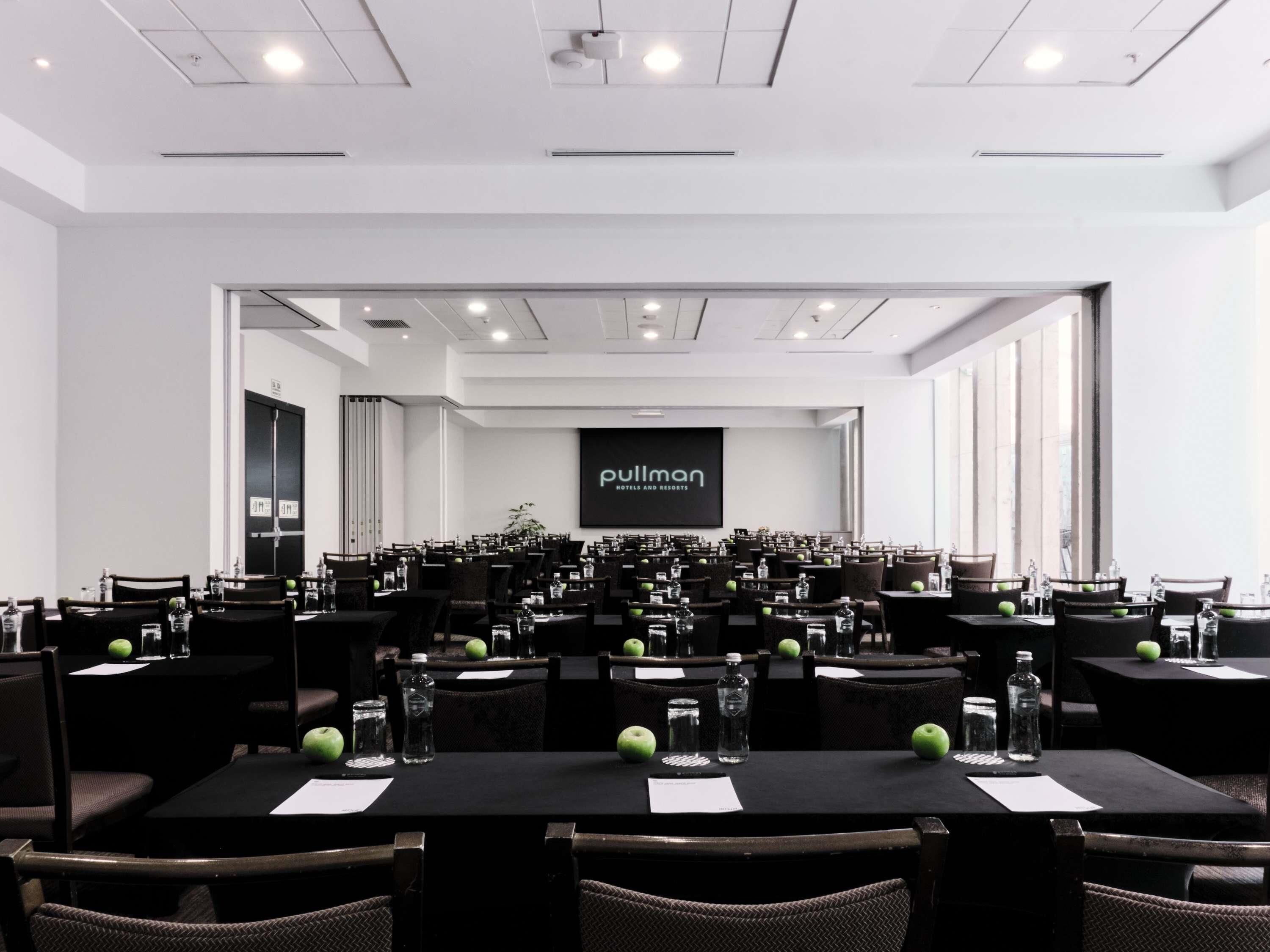A conference room with multiple tables and chairs, a large screen displaying 'Pullman Hotels and Resorts', and a door leading to another room.