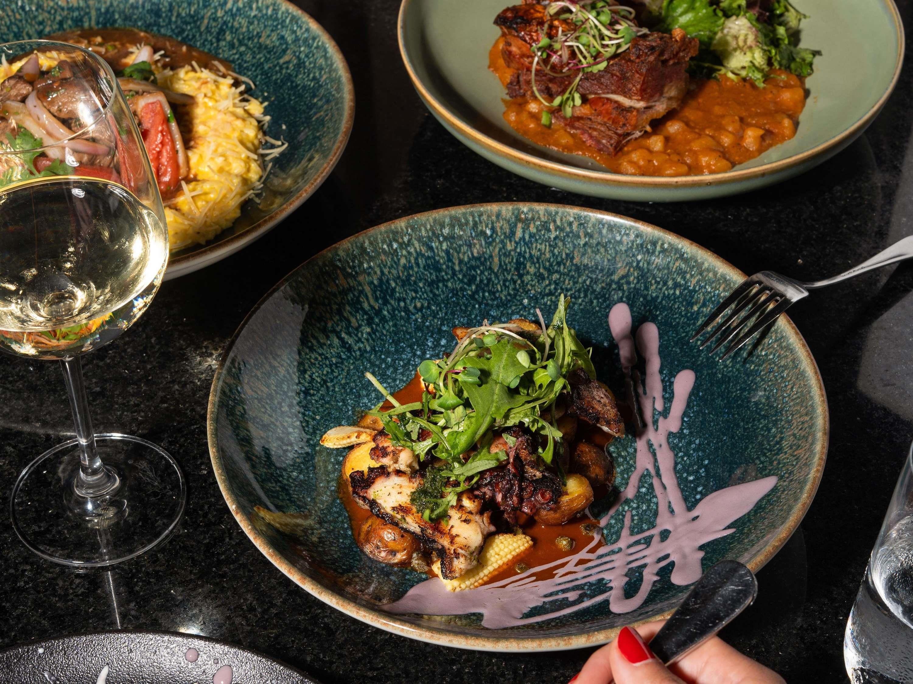 A dining table with three plates of food, a wine glass, and a fork.
