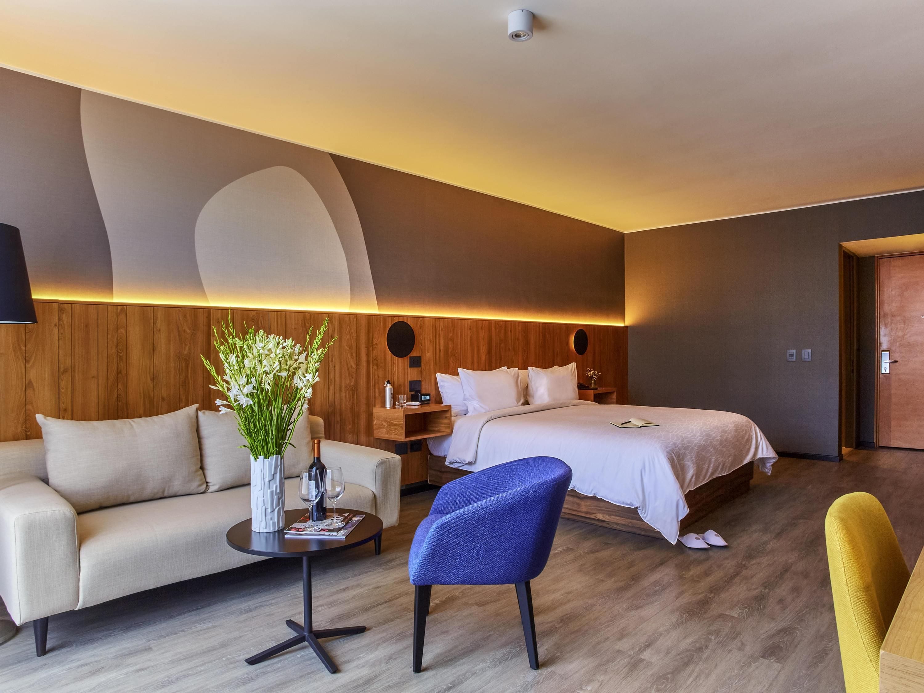 A modern hotel room with a sofa, a small table with a vase of flowers, a blue chair, a bed with white bedding, and a wooden headboard.