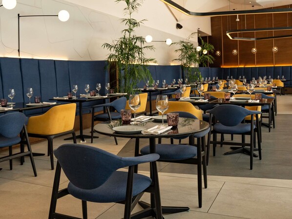 A modern restaurant interior with round tables, blue and yellow chairs, and potted plants.