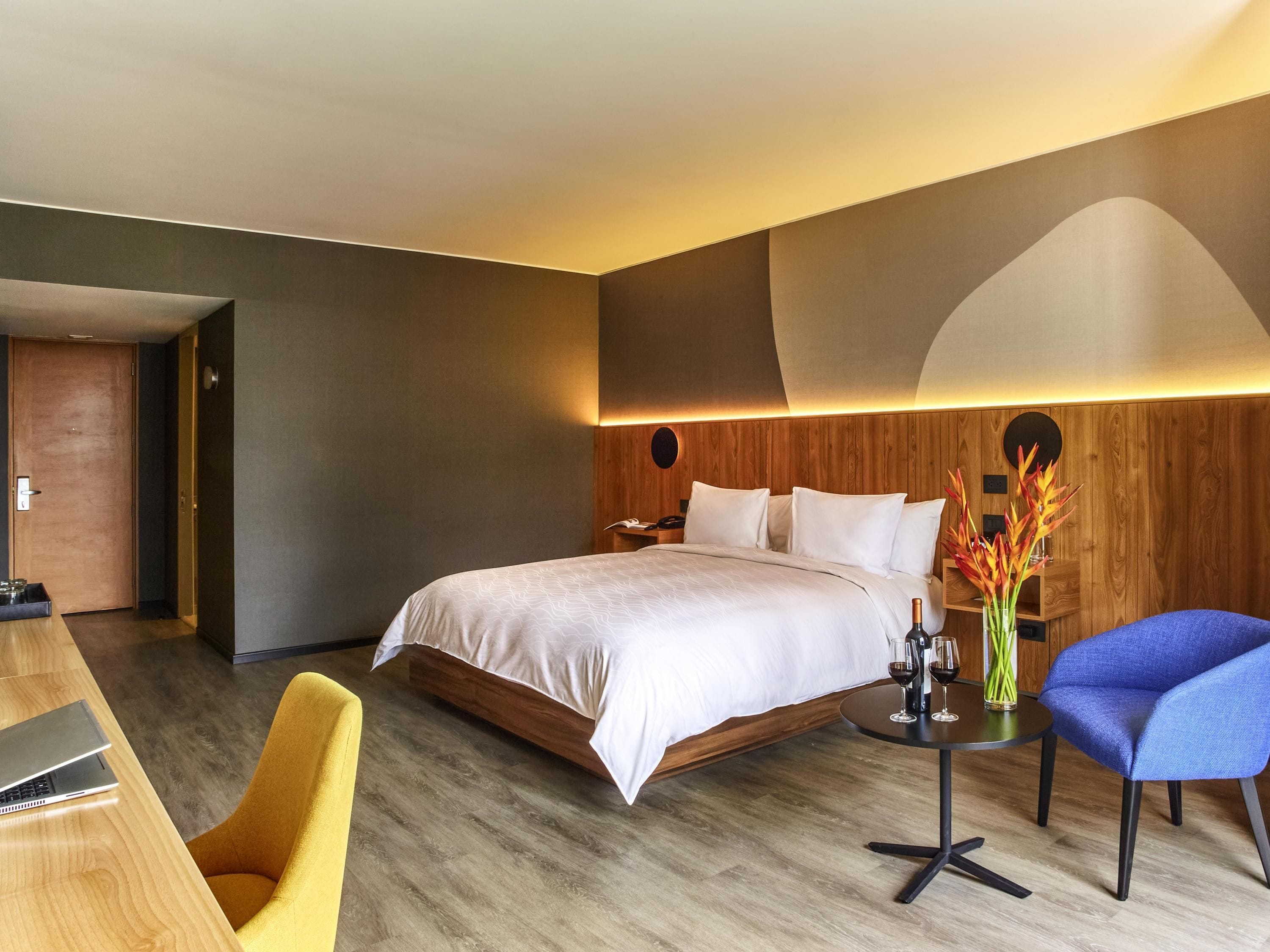 A modern hotel room with a large bed, a wooden headboard, a desk with a laptop, a blue chair, and a vase with flowers.