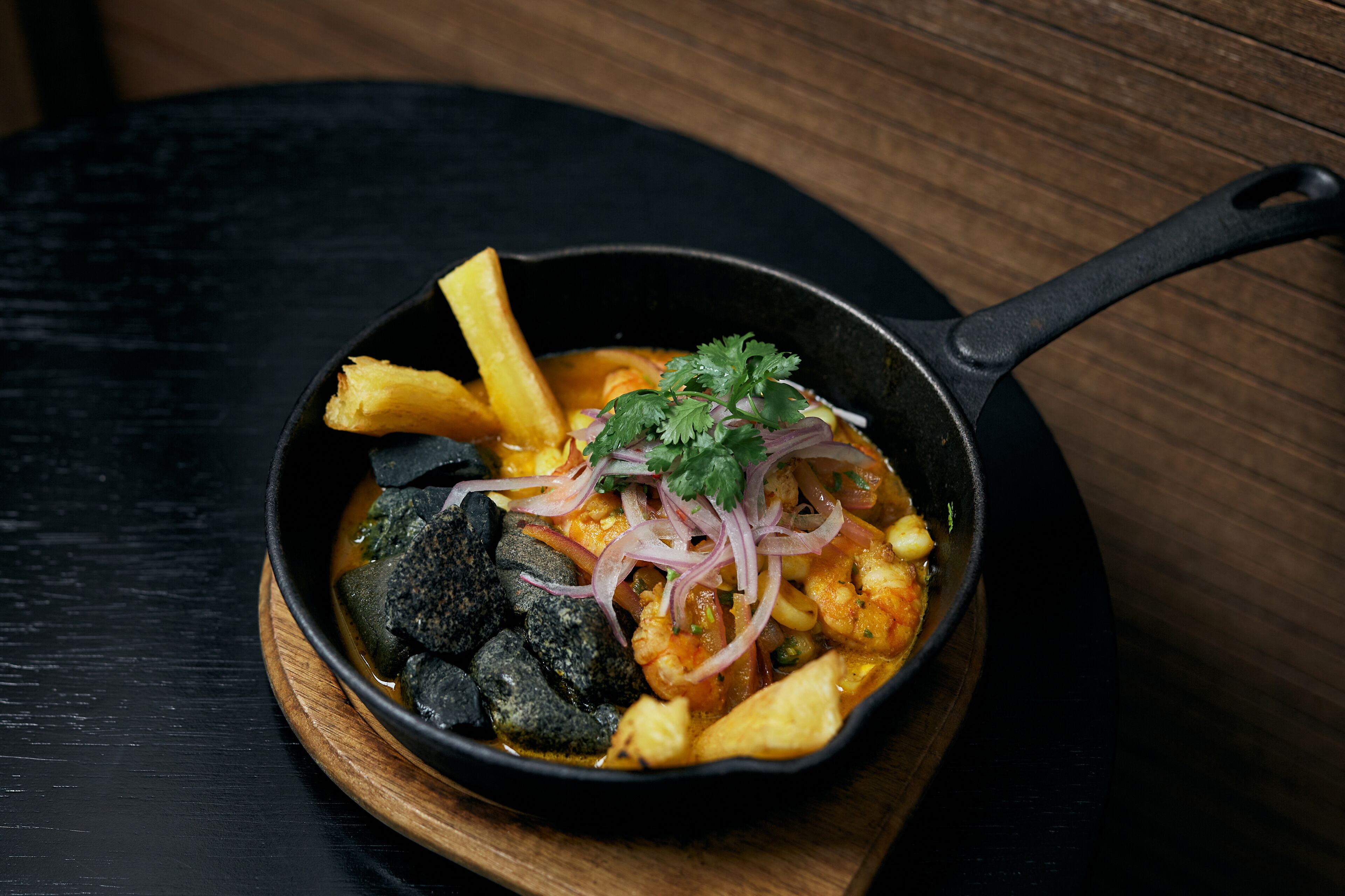 A black skillet with a wooden handle containing a seafood dish, garnished with red onions and cilantro, on a wooden board.