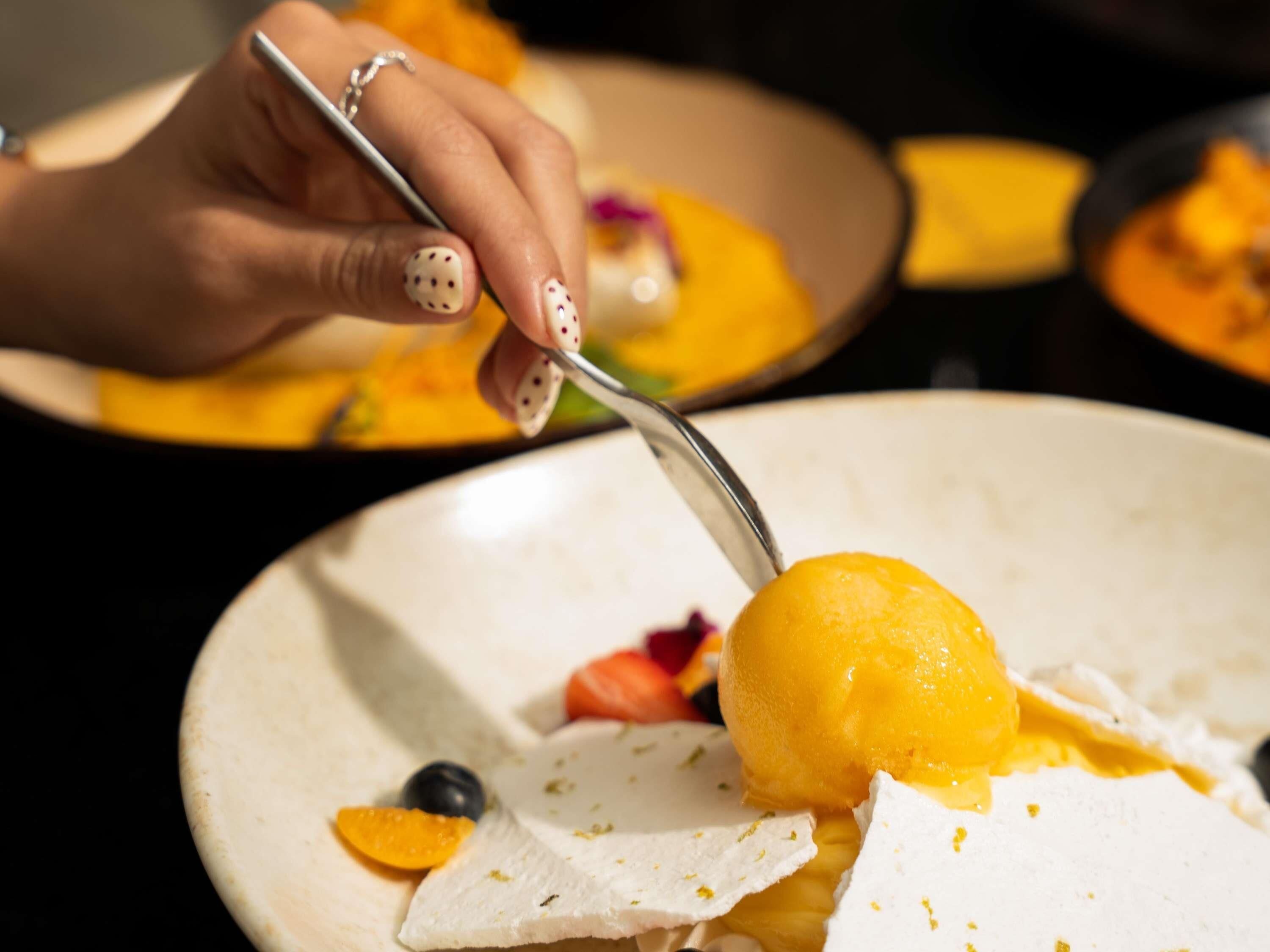 A hand holding a spoon over a plate with a yellow dessert, possibly ice cream, and some fruit pieces.