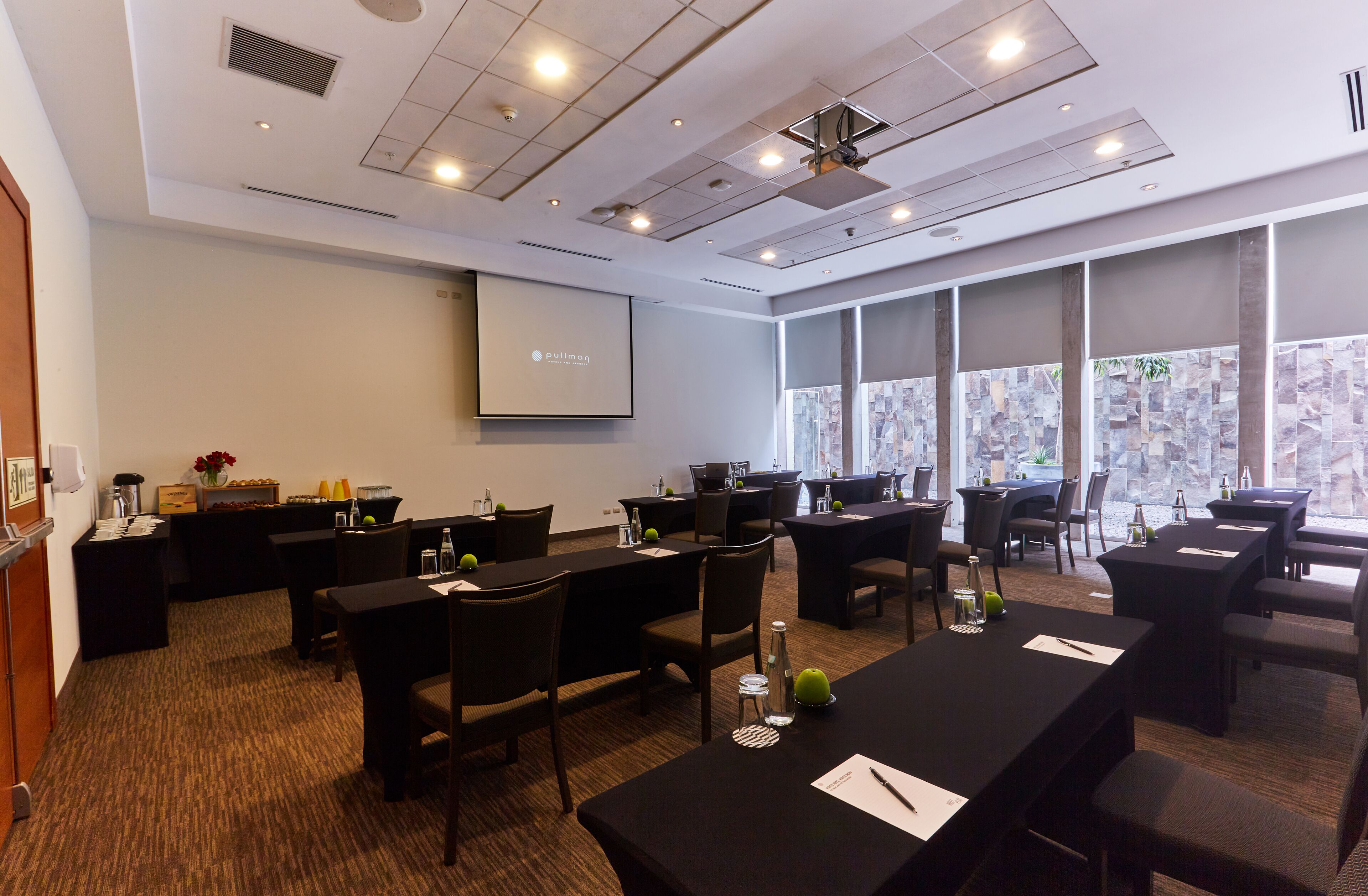 A conference room with multiple tables and chairs, a projector screen, and a large window with a city view.