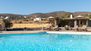 A swimming pool with lounge chairs and umbrellas, surrounded by stone buildings and a view of the countryside. - Swiss Home (Paros)