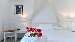 A neatly made bed with white linens, a white chair, a bedside table with a lamp, and a framed picture on the wall. - Swiss Home (Paros)
