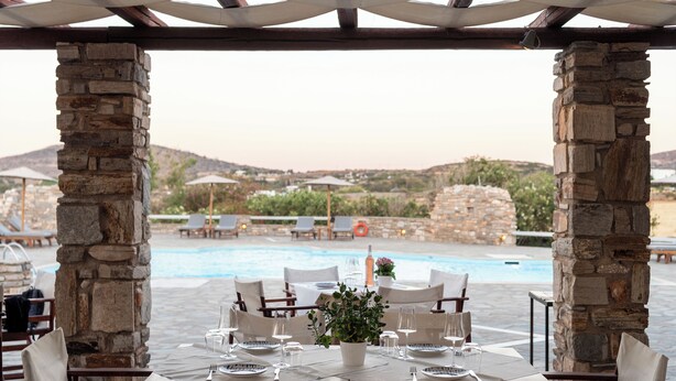 An outdoor dining area with stone pillars, tables set for two, and a view of a swimming pool and hills in the background. - Swiss Home (Paros)
