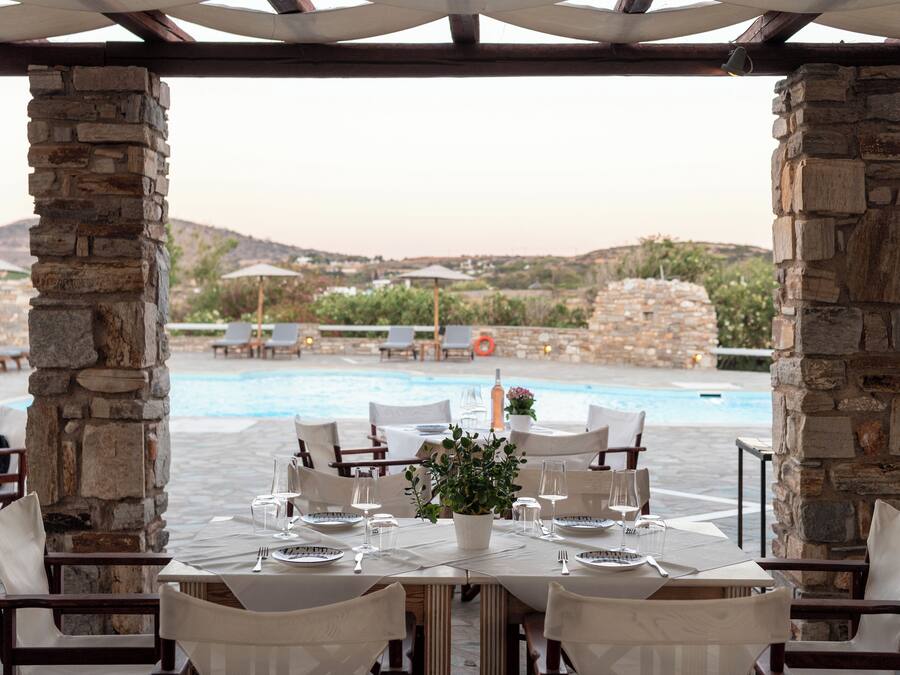 Un área de comedor al aire libre con pilares de piedra, mesas dispuestas para dos personas y vistas a una piscina y colinas al fondo.
