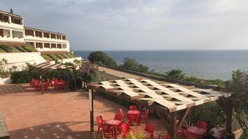 Eine Terrasse mit roten Stühlen und Tischen, die auf das Meer blickt, ein Gebäude mit Balkonen und eine Pergola.