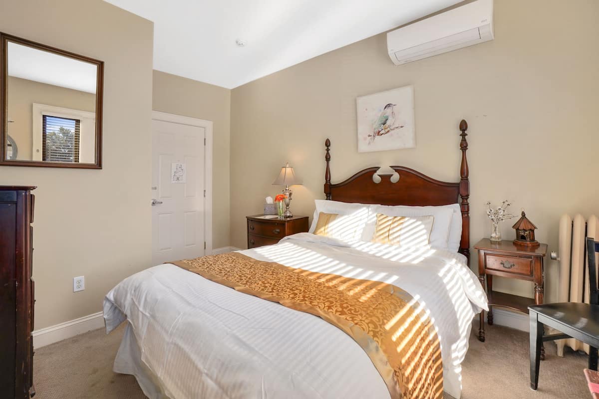 A bedroom with a large bed, a nightstand with a lamp, a dresser, a mirror, and a wall-mounted air conditioner.