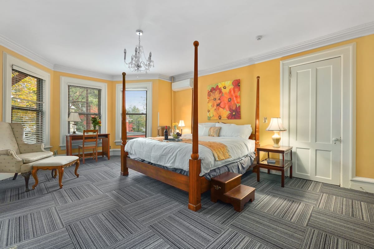 A four-poster bed with a wooden frame, a chandelier, a large floral painting, and a view of the outdoors through a window with blinds.