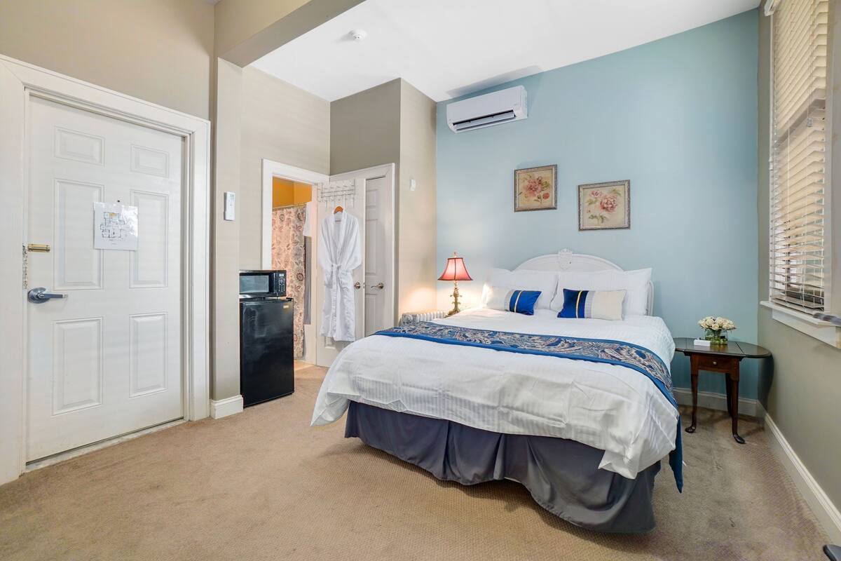 A bedroom with a large bed, a bedside table, an air conditioning unit, and a door with a whiteboard.