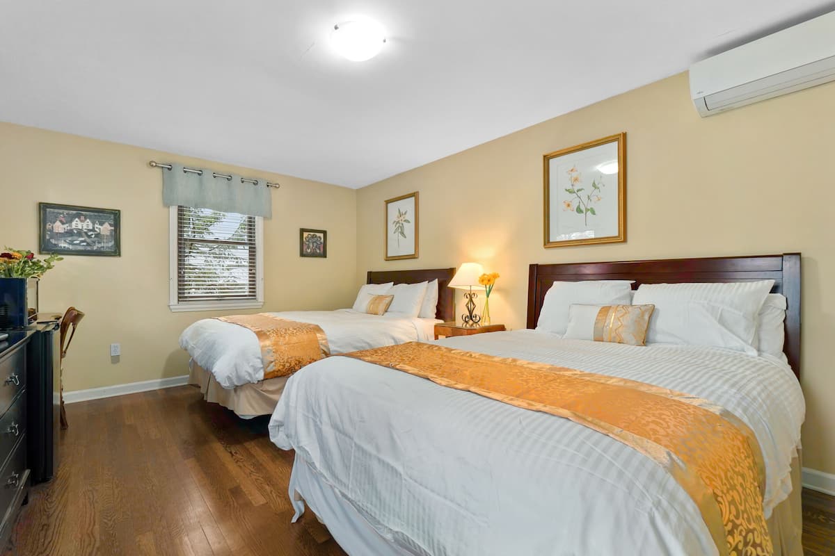 A hotel room with two beds, a dresser, a window with blinds, and wall-mounted air conditioning.