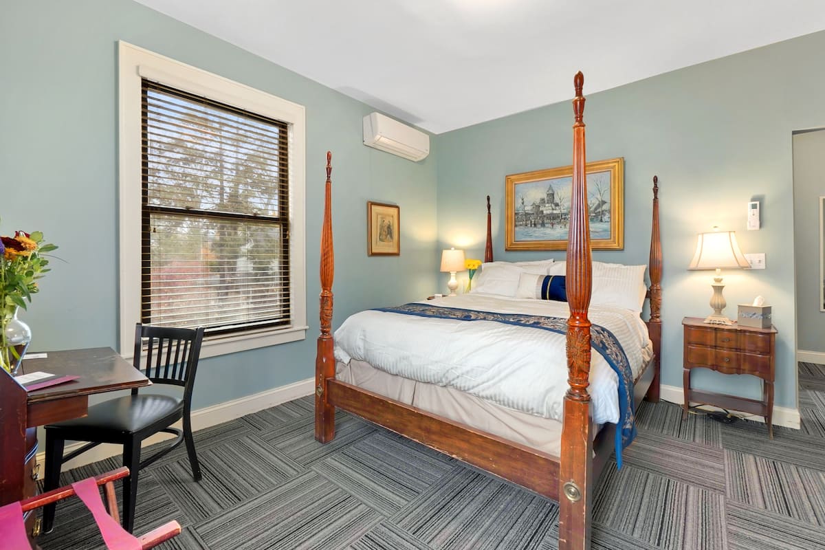 A four-poster bed with a blue and white bedspread, a wooden nightstand with a lamp, a desk with a chair, and a window with blinds.