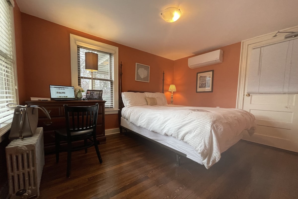 A bedroom with a bed, a desk with a laptop, a chair, a radiator, and a window with blinds.