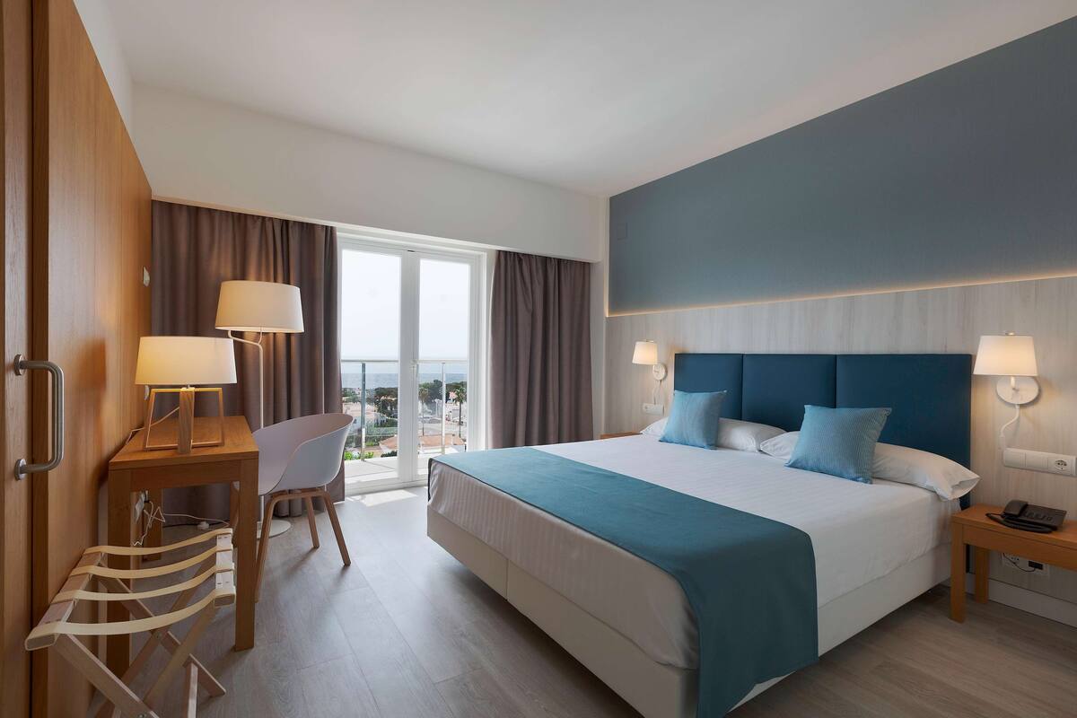 A hotel room with a large bed, two bedside lamps, a desk with a chair, and a view of the outdoors.