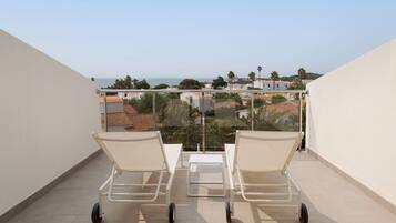A balcony with two white lounge chairs and a small table, overlooking a residential area with houses and palm trees.