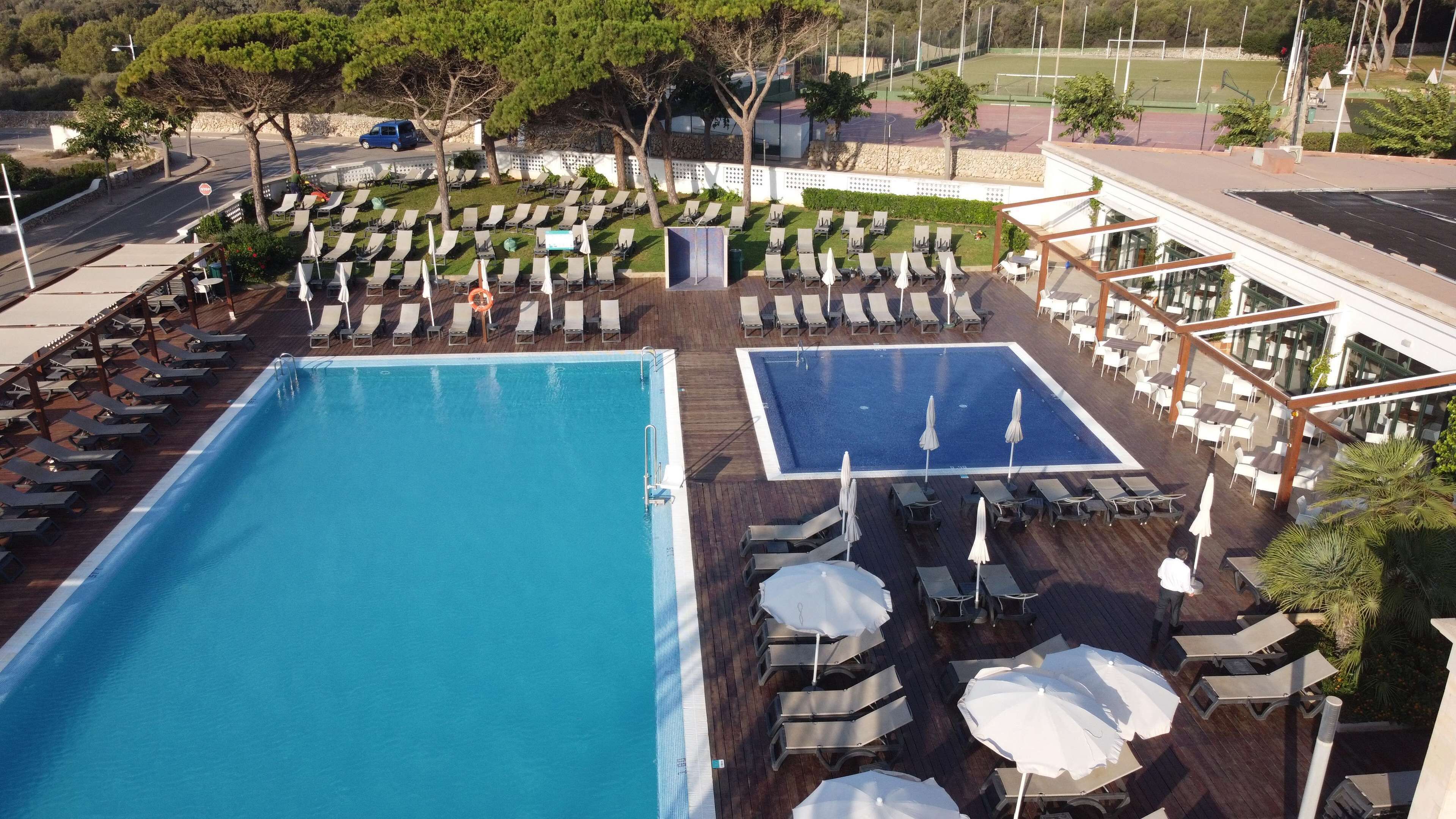 Un espace piscine aménagé sur le toit, doté de plusieurs chaises longues et de parasols, entouré d’une terrasse en bois et offrant une vue sur un court de tennis.