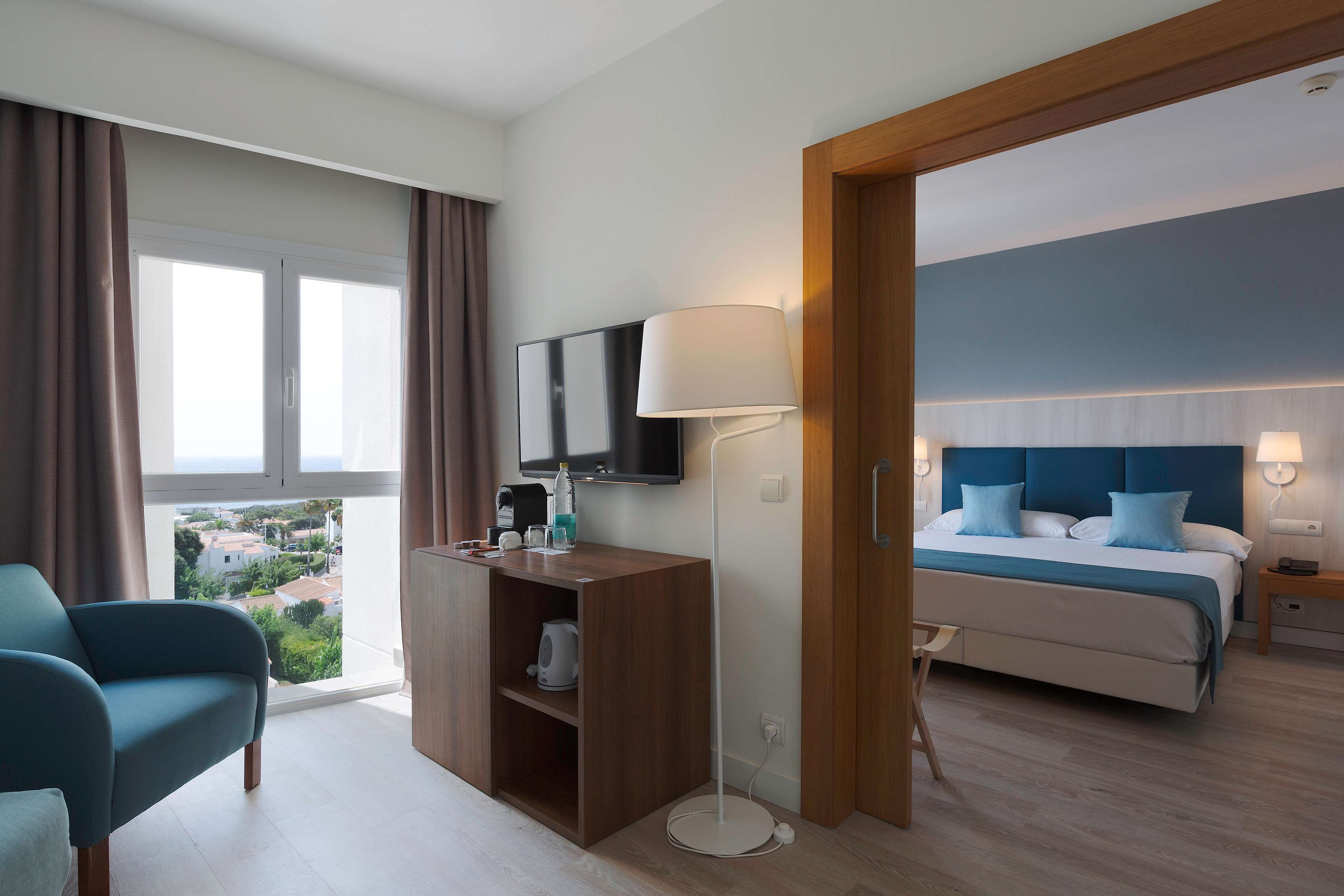 A hotel room with a bed, a desk with a flat-screen TV, a lamp, and a view of the city.
