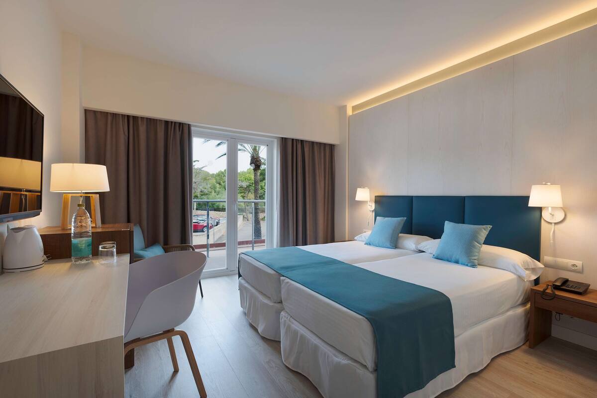 A hotel room with a bed, two bedside tables, a desk with a chair, a window with curtains, and a view of the outdoors.