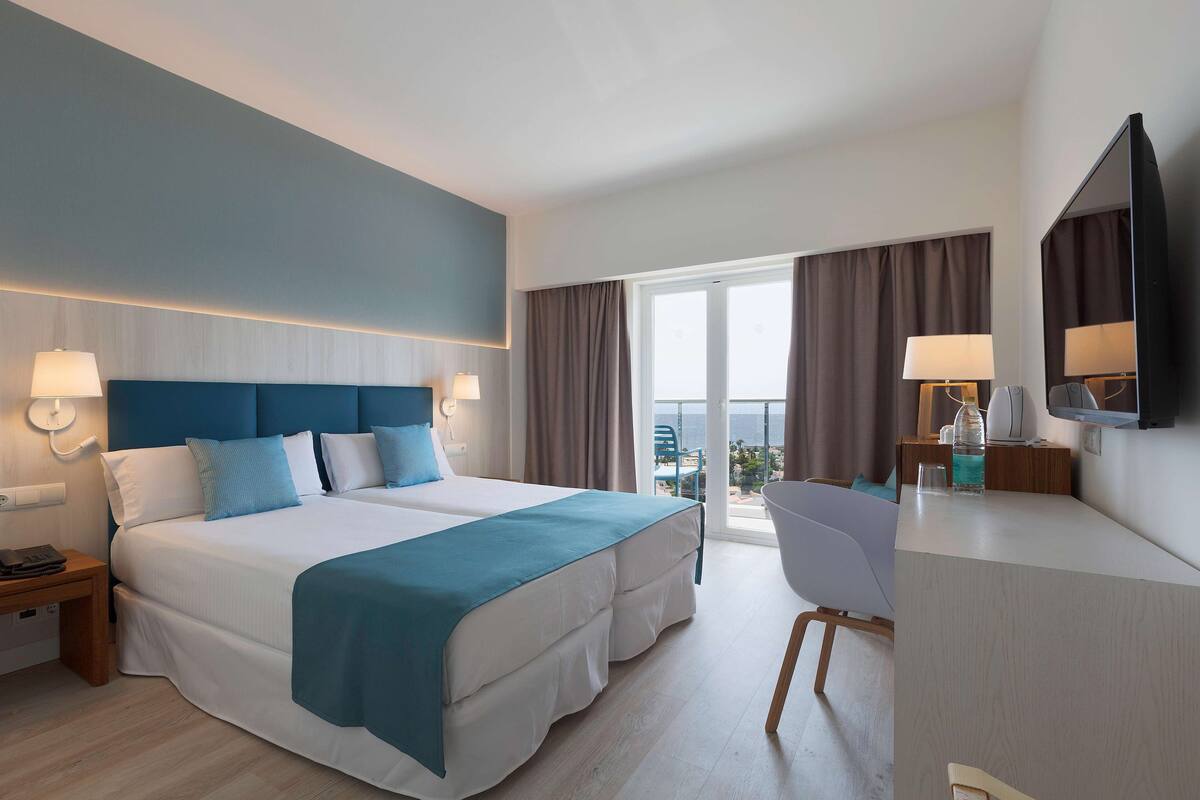 A hotel room with a large bed, a desk, a chair, a TV, and a balcony with curtains.