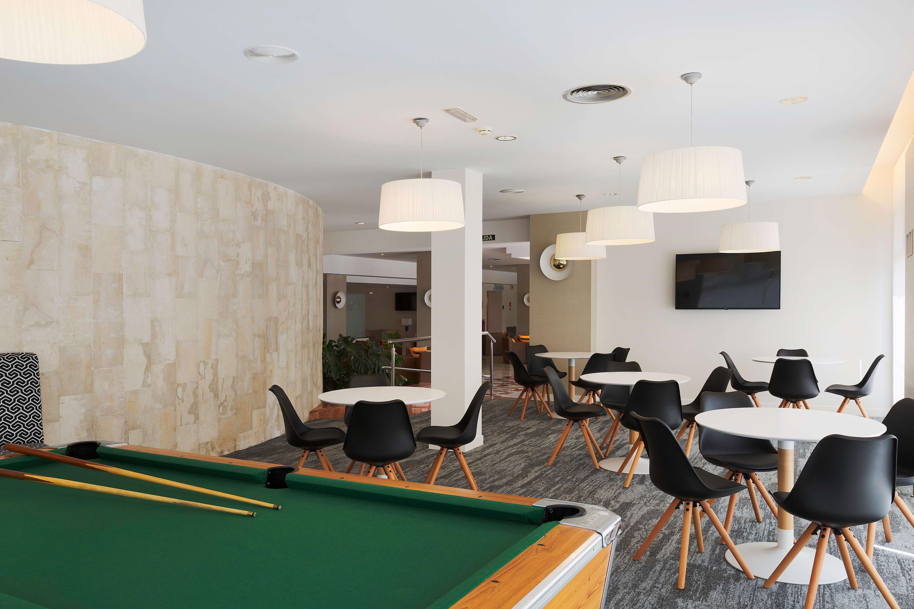 A pool table with black chairs and a green felt surface, a white table with black chairs, and a large flat-screen TV mounted on the wall.