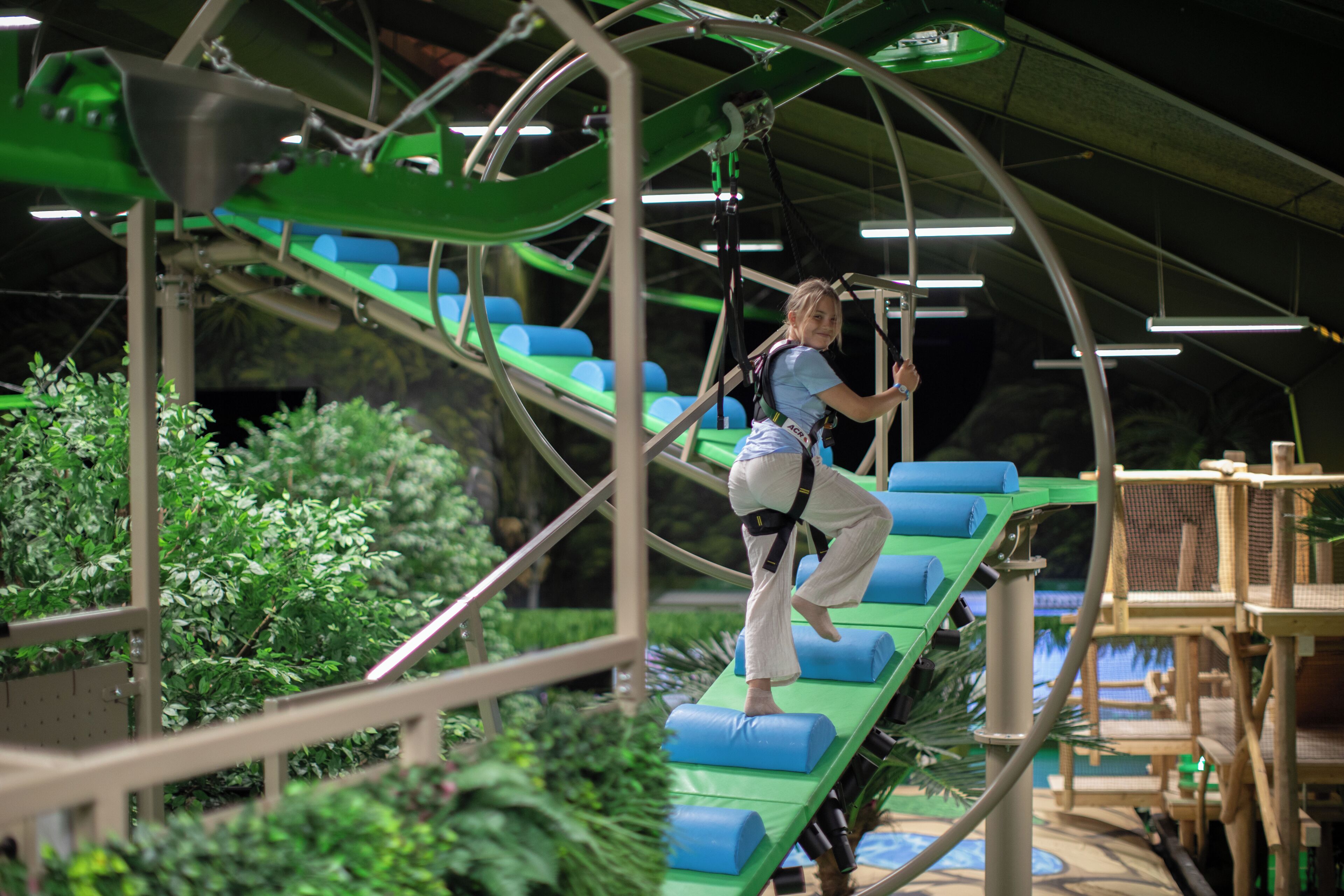 Una persona en un circuito de tirolesa con escalones acolchados de color verde y azul, rodeada de follaje y estructuras metálicas.