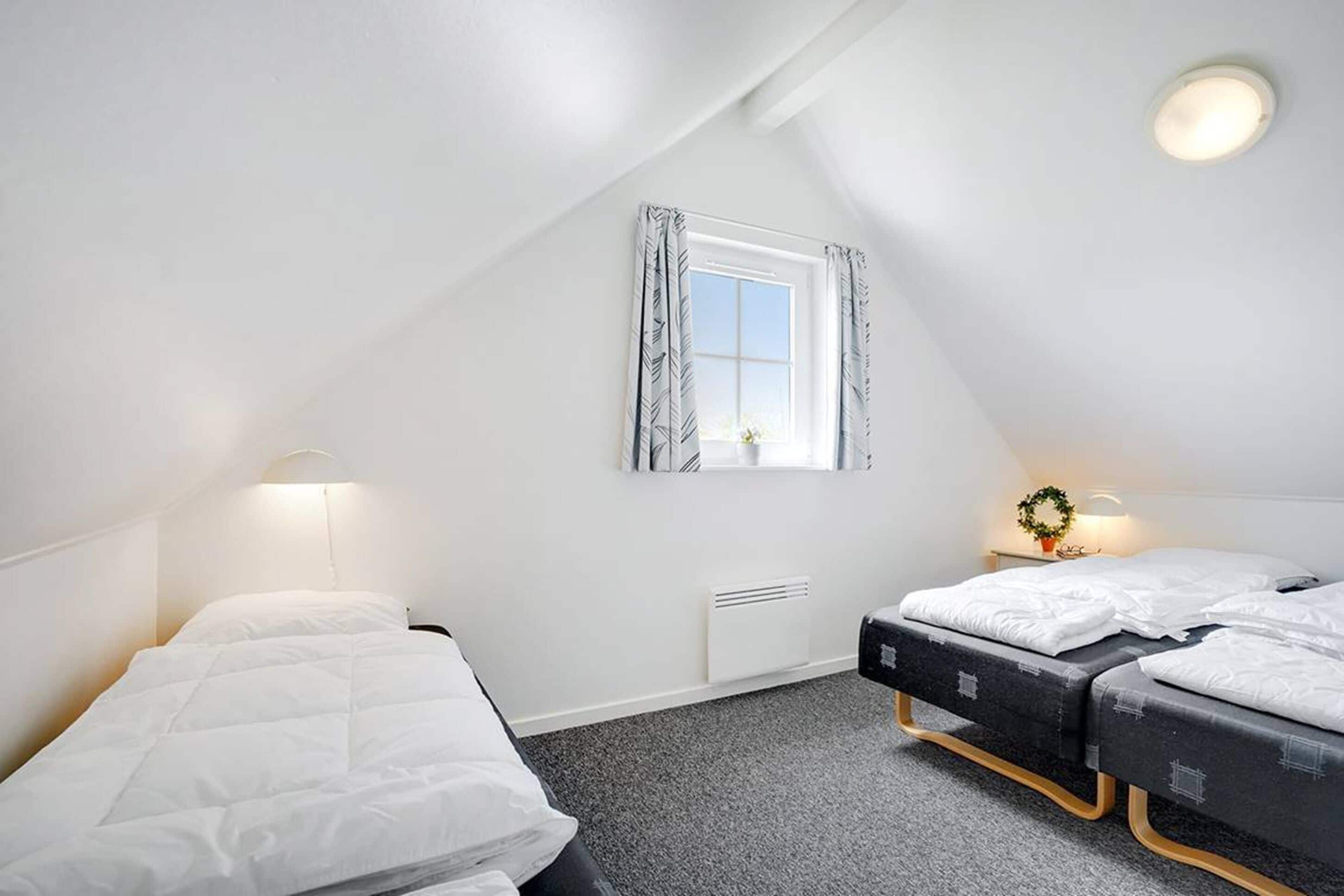 Une petite pièce bien éclairée, avec deux lits, une fenêtre avec des rideaux, un radiateur et un luminaire fixé au mur.
