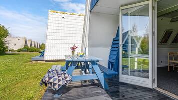 Eine Terrasse mit Holztisch und -stühlen, ein kleiner Garten und ein Gebäude mit weißer Fassade und Glastür.