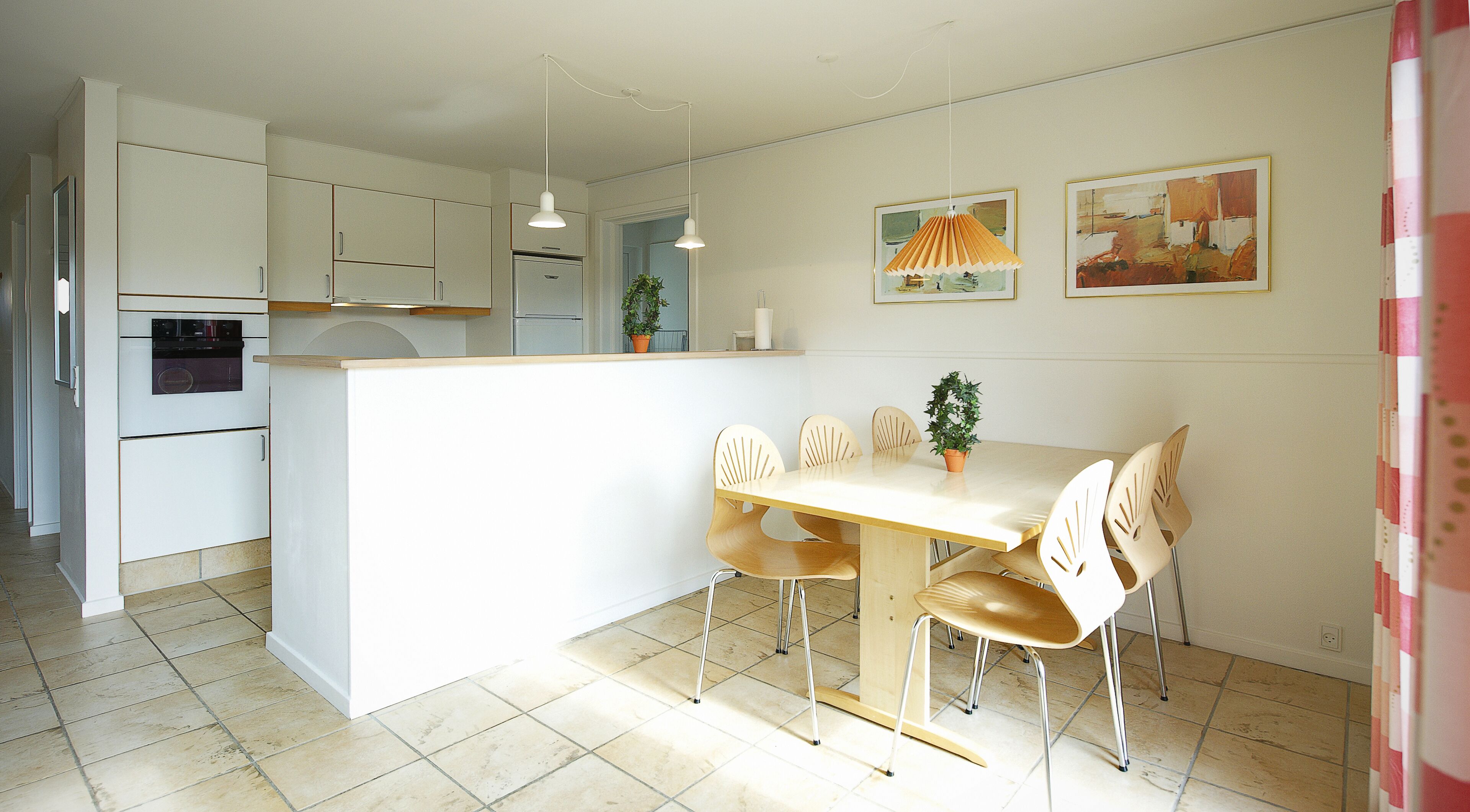 A modern kitchen with white cabinets, a built-in oven, and a dining area with a wooden table and chairs. There are two pendant lights above the table and a potted plant on the counter.