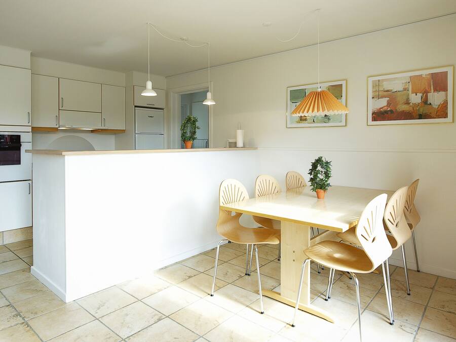 A modern kitchen with white cabinets, a built-in oven, and a dining area with a wooden table and chairs. There are two pendant lights above the table and a potted plant on the counter.