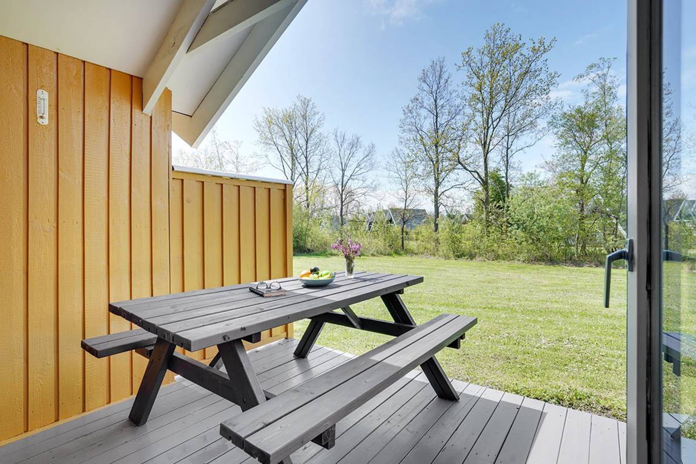 Una terraza de madera con una mesa de picnic, un ventanal de vidrio y vistas a un área verde con árboles.