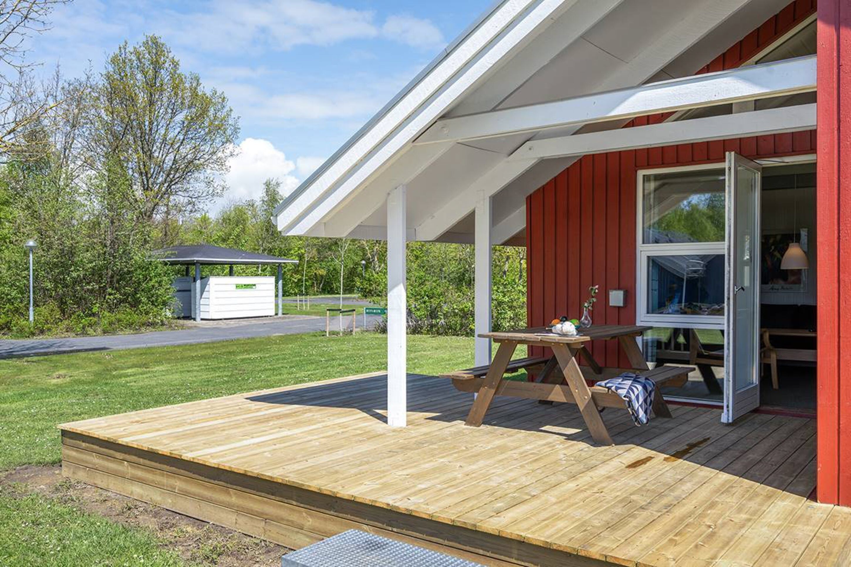 Eine Holzterrasse mit Picknicktisch und Bänken, einem überdachten Sitzbereich und einem roten Gebäude mit Glastür.