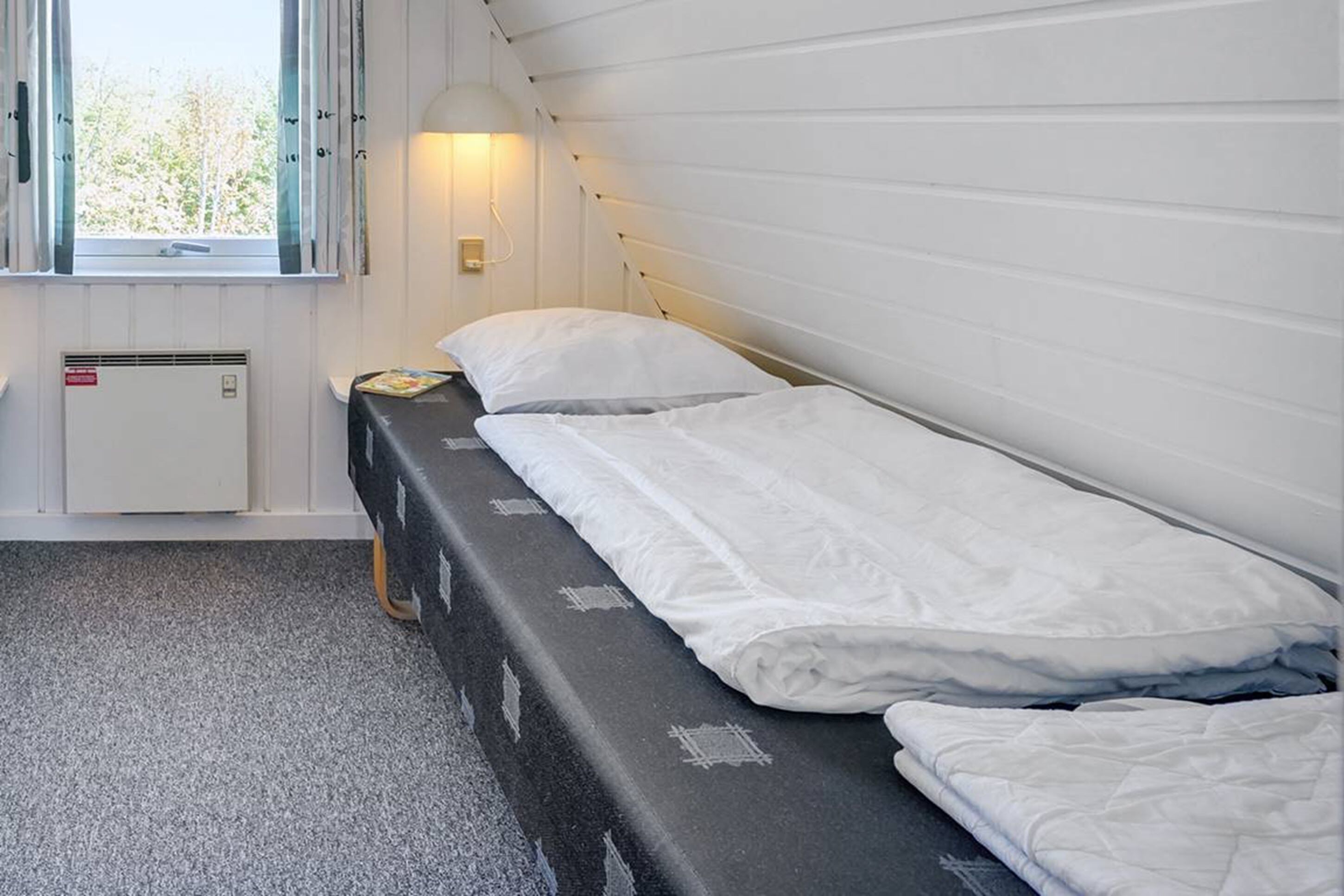 Ein Einzelbett mit weißer Bettwäsche, ein Nachttisch mit Lampe und ein Fenster mit Vorhängen.