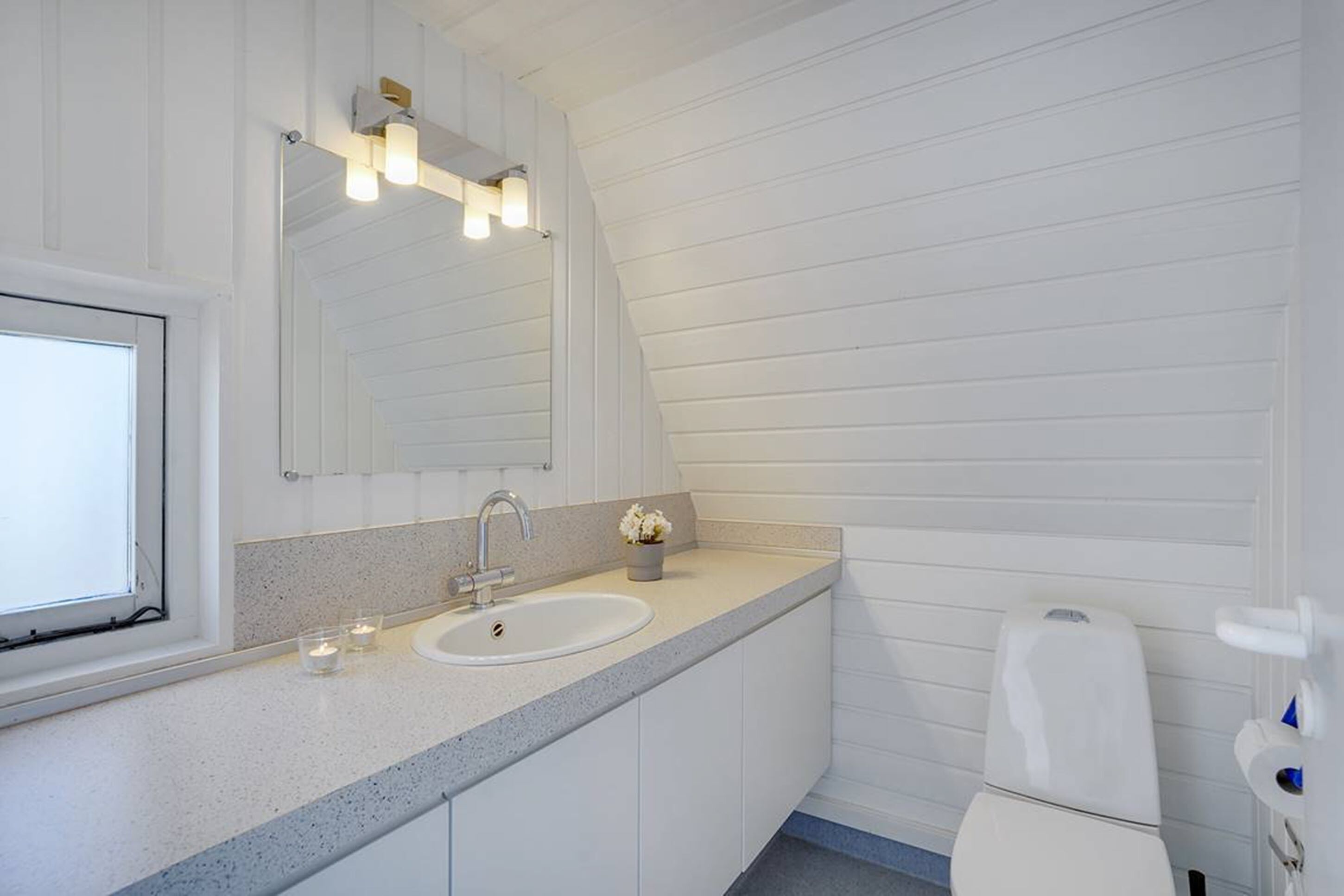 Ein Badezimmer mit weißem Waschbecken, Spiegel und Toilette. Links befindet sich ein Fenster und über dem Spiegel eine Leuchte.