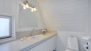 Ein Badezimmer mit weißem Waschbecken, Spiegel und Toilette. Links befindet sich ein Fenster und über dem Spiegel eine Leuchte.